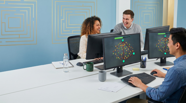 Drei Mitarbeitende spielen einen online escape room, sie sitzen an Computern in einem Büro und lächeln sich gegenseitig an.